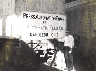 AFCO-Press-Automation-Equip-being-shipped-1 Black-and-white historical photo of a man standing beside a truck loaded with industrial equipment, with a sign reading “Press Automation Equip. by Automatic Feed Co., Napoleon, Ohio” displayed on the shipment.