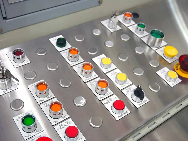 Close-up of a stainless steel industrial control panel featuring multiple illuminated indicator lights in red, orange, yellow, green, and white. The panel includes push buttons, selector switches, a key-operated control switch, and a large red emergency stop button on the right. Labels beneath the controls identify various machine functions and feed system settings.